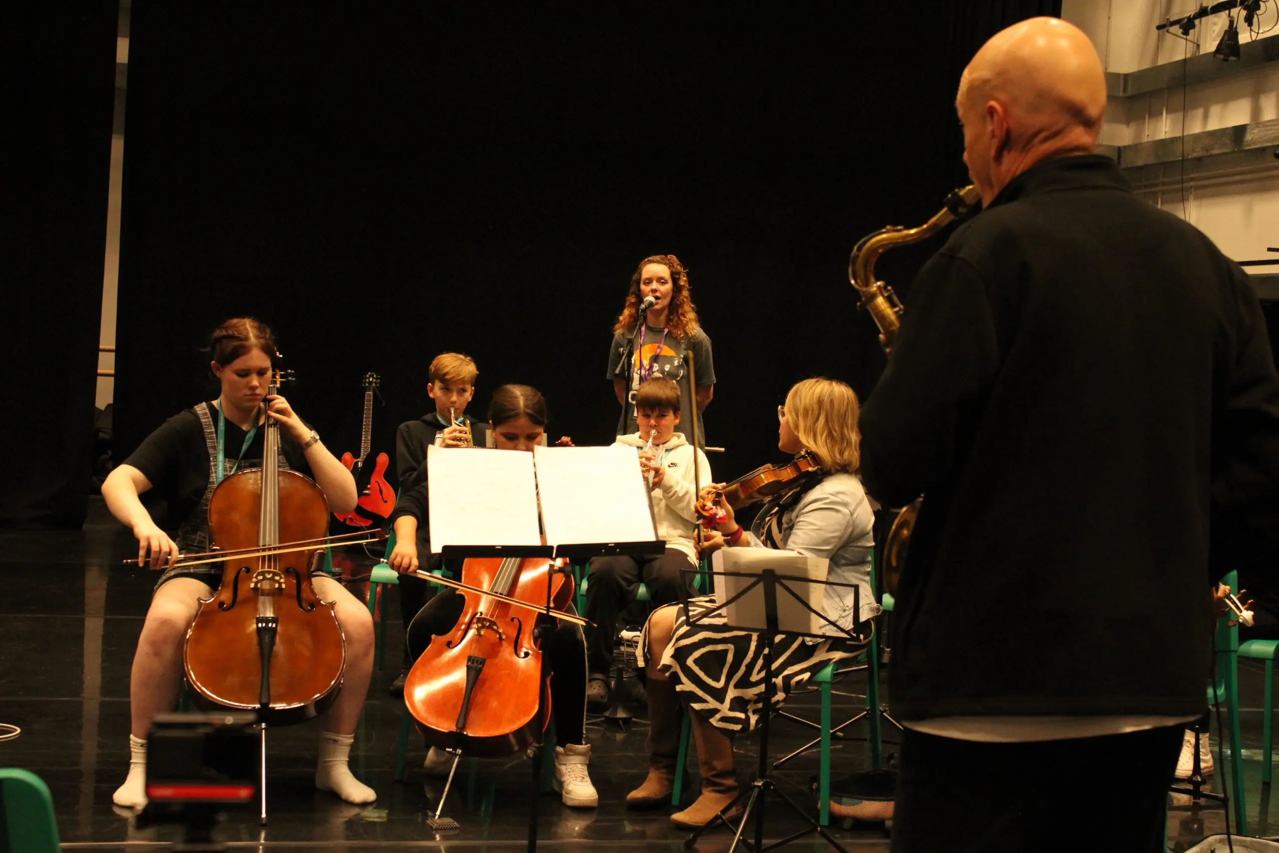 Young musicians in an ensemble playing various instruments including cellos, violin, saxophone, trumpet and vocals.