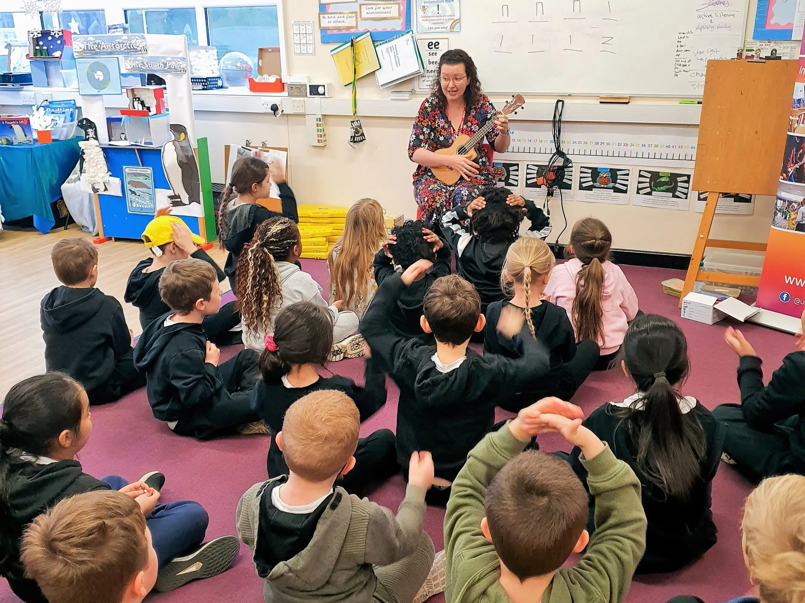 A music teacher holding a ukulele sits on a chair in front of a class of primary school children who are sat on the carpet
