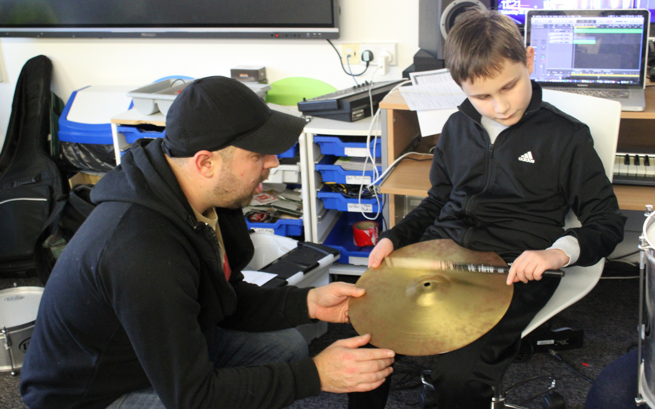 A white man in a black cap is crouched next to a young blind white boy in a black jacket who is interacting with a cymbal, offering support in the boy's musical journey