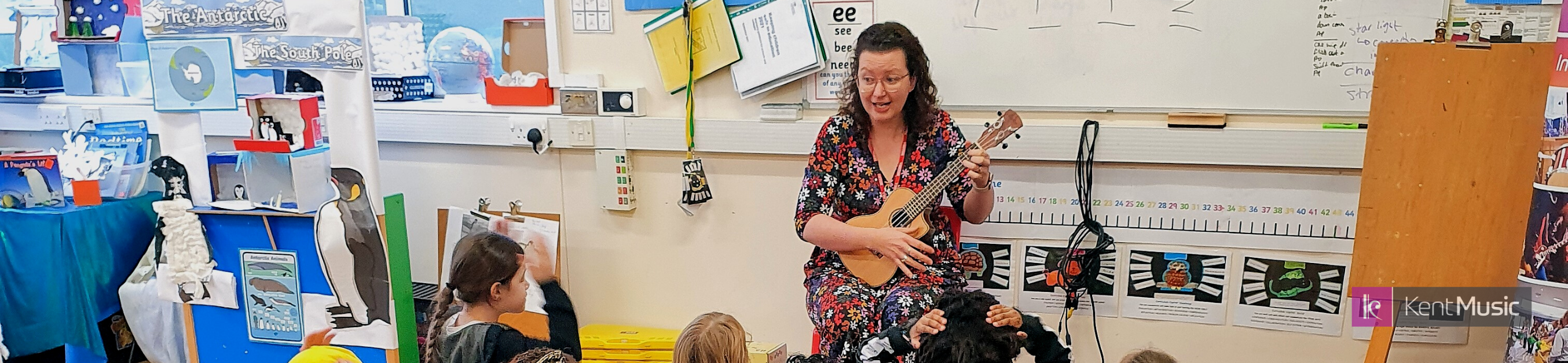 a woman plays the ukulele and sings in front of a group of seated primary aged children