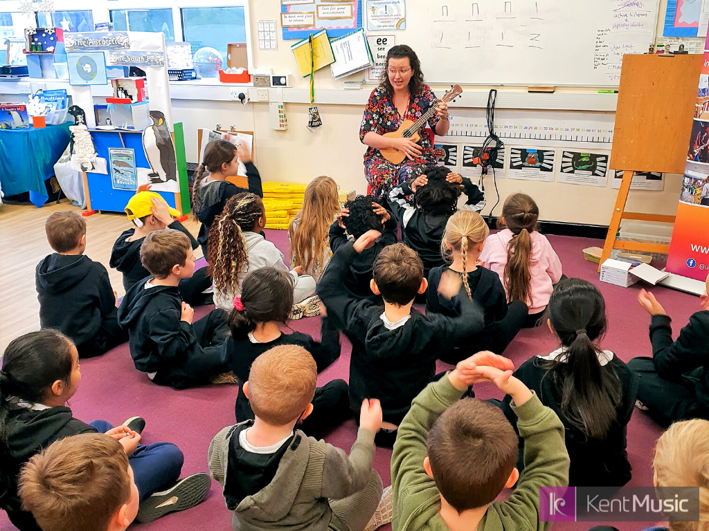 a woman plays the ukulele and sings in front of a group of seated primary school aged children in a classroom setting